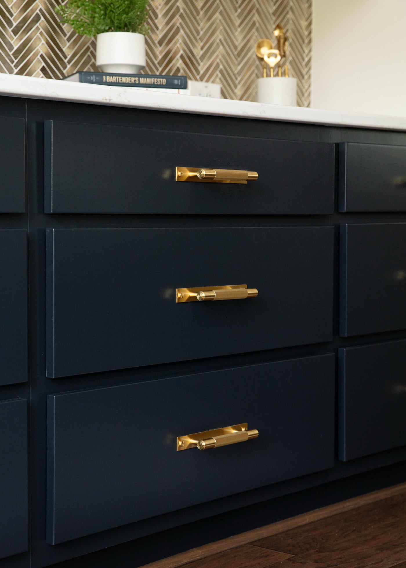Navy blue cabinet drawers with long faceted brass bar pulls beneath a white stone counter in the Vue Point project by Jennifer Kizzee Design